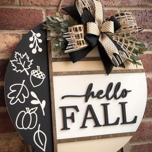 May include: A round wooden door hanger with a black and white striped bow, greenery, and the words "hello fall" in black lettering.