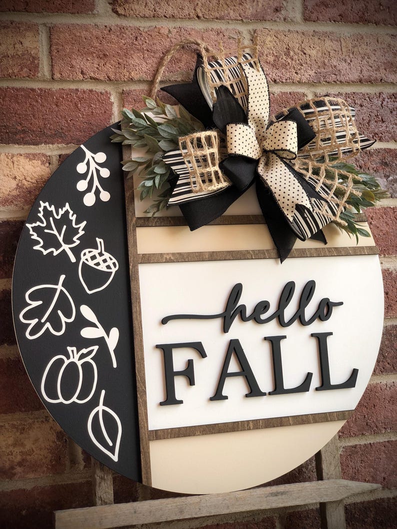 May include: A round wooden door hanger with a black and white striped bow, greenery, and the words "hello fall" in black lettering.
