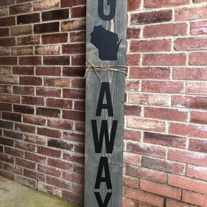 May include: A wooden sign with the word "G" and the outline of the state of Wisconsin, followed by the word "AWAY" spelled vertically. The sign is painted gray with black lettering.