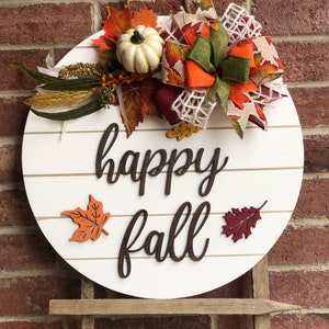 May include: A white round wooden sign with the words "happy fall" in brown lettering. The sign is decorated with fall-themed embellishments, including a small white pumpkin, orange and brown leaves, and a burgundy and green bow.
