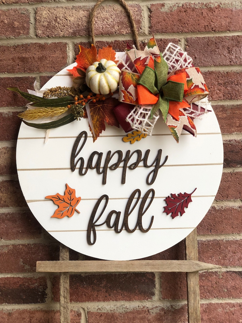 May include: A white round wooden sign with the words "happy fall" in brown lettering. The sign is decorated with fall-themed embellishments, including a small white pumpkin, orange and brown leaves, and a burgundy and green bow.