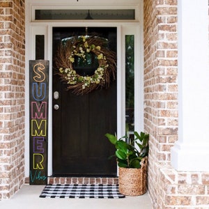 May include: A wooden sign with the word "SUMMER" in colorful neon letters and the word "Vibes" in cursive. The sign is placed on a porch with a black door, a wreath, and a black and white checkered mat.