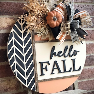 May include: A round wooden door hanger with a black and white chevron pattern, a peach colored background, and the words "hello fall" in black lettering. The hanger is decorated with a small orange pumpkin, burlap, and black and white ribbon.