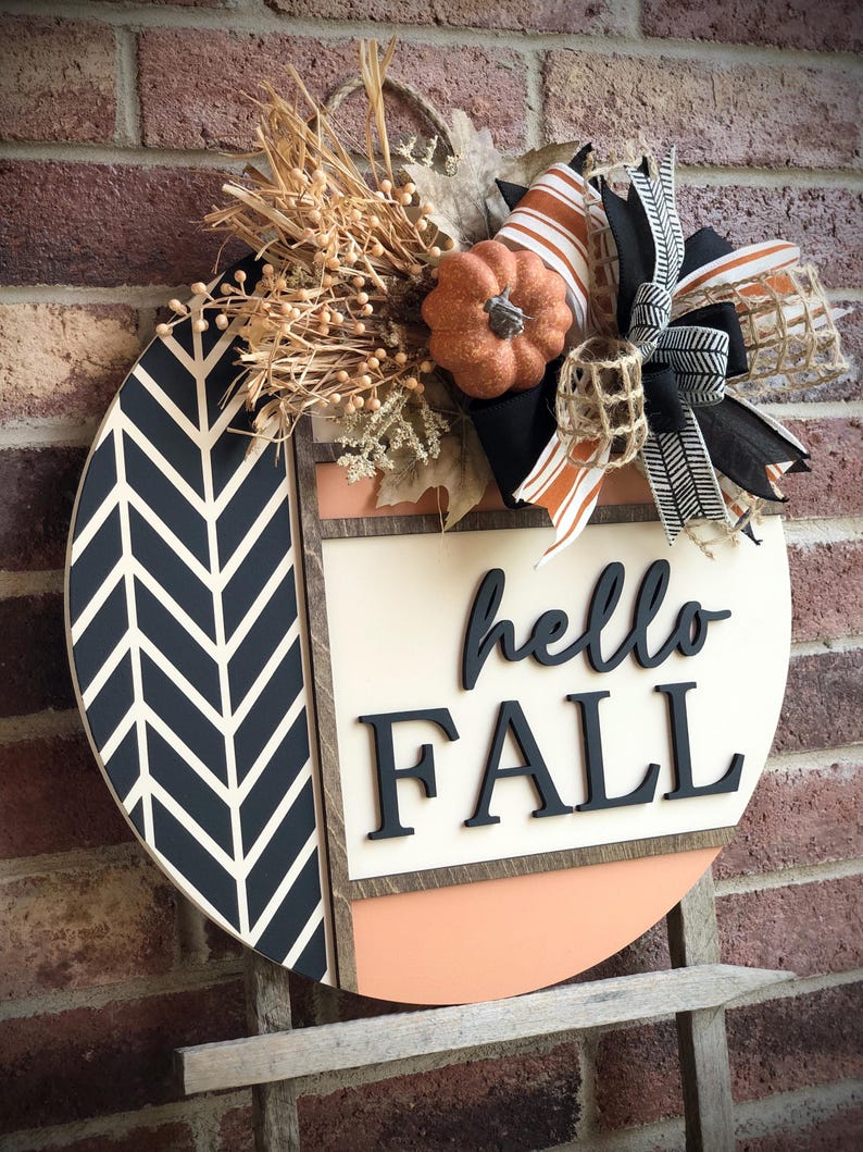 May include: A round wooden door hanger with a black and white chevron pattern, a peach colored background, and the words "hello fall" in black lettering. The hanger is decorated with a small orange pumpkin, burlap, and black and white ribbon.