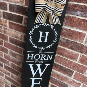 May include: Black wooden welcome sign with a white wreath and the letter H in the center. The sign also says "The Horn Family" and "Welcome" in white letters.