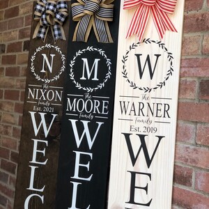 May include: Three wooden welcome signs with different family names and established dates. The signs are painted in black, white, and natural wood. Each sign has a wreath with a letter in the center and a bow at the top.