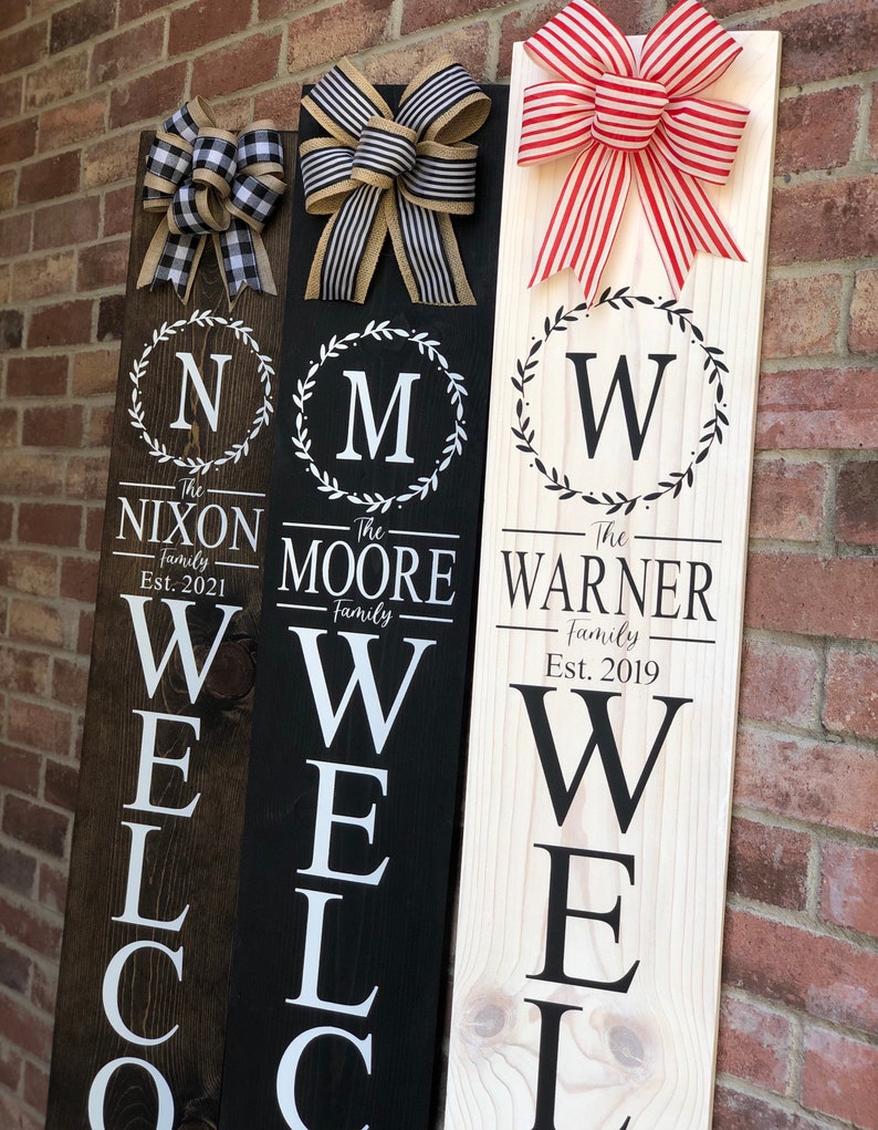 May include: Three wooden welcome signs with different family names and established dates. The signs are painted in black, white, and natural wood. Each sign has a wreath with a letter in the center and a bow at the top.