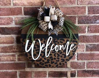 Puede incluir: Un colgador de puerta redondo de madera con un diseño de estampado de leopardo y la palabra "Bienvenido" en letras blancas. El colgador está decorado con un lazo negro, blanco y marrón.