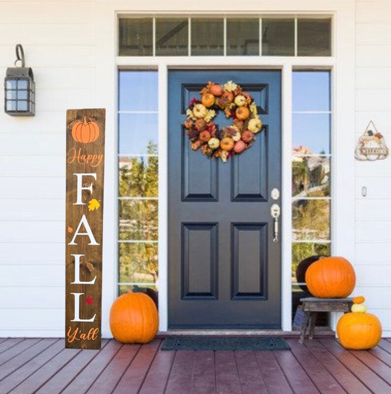 May include: A wooden sign with the text "Happy Fall Y'all" and a pumpkin graphic. The sign is leaning against a white door with a fall wreath. There are pumpkins on the porch.
