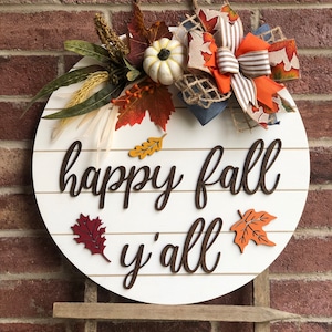 Puede incluir: Un letrero redondo de madera blanca con las palabras "happy fall y'all" en escritura marr&oacute;n. El letrero est&aacute; decorado con una corona de oto&ntilde;o con hojas naranjas, rojas y marrones, una peque&ntilde;a calabaza blanca y un lazo marr&oacute;n y blanco a rayas.