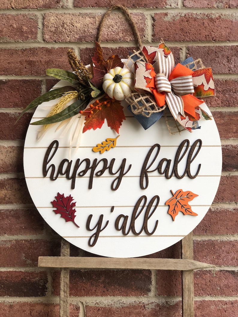 Puede incluir: Un letrero redondo de madera blanca con las palabras "happy fall y'all" en escritura marr&oacute;n. El letrero est&aacute; decorado con una corona de oto&ntilde;o con hojas naranjas, rojas y marrones, una peque&ntilde;a calabaza blanca y un lazo marr&oacute;n y blanco a rayas.