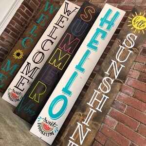 May include: A collection of wooden welcome signs with various summer-themed phrases and graphics. The signs are rectangular and feature text like "Welcome," "Summer," and "Sunshine." Each sign has a unique design, including a sunflower, watermelon, and sun.