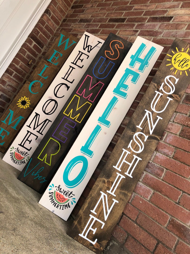 May include: A collection of wooden welcome signs with various summer-themed phrases and graphics. The signs are rectangular and feature text like "Welcome," "Summer," and "Sunshine." Each sign has a unique design, including a sunflower, watermelon, and sun.