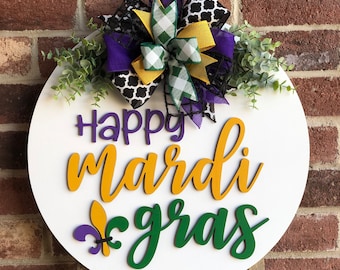 May include: A white round wooden door hanger with the text "Happy Mardi Gras" in gold, green, and purple lettering. A purple, green, and gold bow with black and white checkered ribbon is attached to the top.