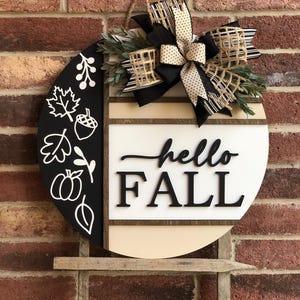 May include: A round wooden door hanger with a black and white design. The left side features a black background with white fall-themed illustrations, including leaves, acorns, and pumpkins. The right side features a white background with the text "hello FALL" in black. A black and white bow with a burlap accent is attached to the top.