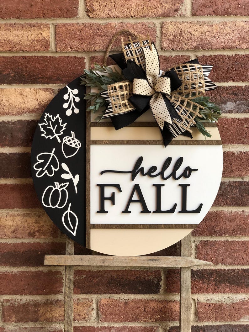 May include: A round wooden door hanger with a black and white design. The left side features a black background with white fall-themed illustrations, including leaves, acorns, and pumpkins. The right side features a white background with the text "hello FALL" in black. A black and white bow with a burlap accent is attached to the top.
