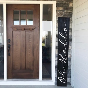 May include: Black wooden sign with white cursive lettering that says "Oh, Hello". The sign is leaning against a brick wall next to a brown wooden door.