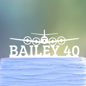 Könnte beinhalten: Ein hellblauer Kuchen mit weißem Flugzeug-Tortentopper mit der Aufschrift "BAILEY 40". Der Kuchen hat eine strukturierte, geriffelte Oberfläche und steht auf einem silbernen Kuchenbrett. Der Hintergrund ist verschwommen.
