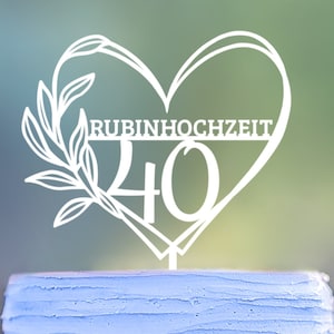 Può includere: Topper per torta bianco a forma di cuore con le parole "Rubinhochzeit 40" all'interno. Il topper per torta è sopra una torta blu chiaro.