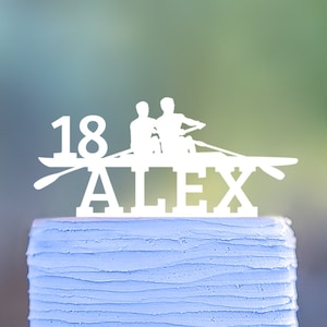 Peut inclure: Un topper de gâteau blanc avec deux personnages en train d'aviron. Le topper indique "18 ALEX". Le gâteau est bleu clair avec un glaçage texturé.