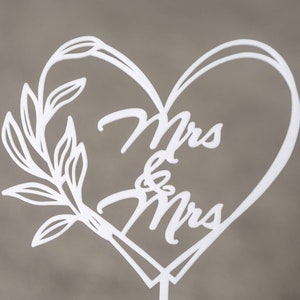 Op de afbeelding: Witte lasergesneden taarttopper met de woorden "Mrs & Mrs" in een hartvormig ontwerp. Een decoratieve bladertak bevindt zich aan de linkerkant van het hart. De topper is gemonteerd op een dunne, witte stok.