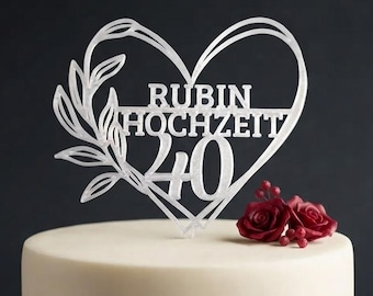 May include: A silver-coloured cake topper shaped like a heart with a leafy branch on the left side. The text RUBIN HOCHZEIT and the number 40 are centred within the heart. The topper is placed on a smooth, round, off-white cake with red roses.