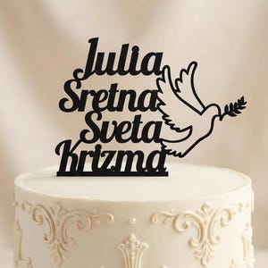 May include: A black cake topper featuring the text Julia Sretna Sveta Krizma and a dove holding an olive branch, positioned on a white cake decorated with cream-colored piped cross motifs and small pearl accents on the sides.