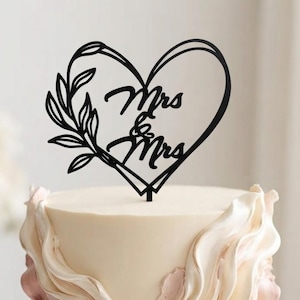 May include: A round cake with smooth cream-colored frosting and decorative vertical ruffles in shades of white and dusty rose. A black cake topper shaped like a heart with a leafy branch on the left side features the text Mrs & Mrs in script.