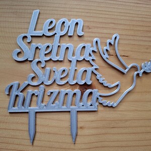 May include: A silver-colored cake topper featuring the text Leon Sretna Sveta Krizma in a cursive font, accompanied by a dove silhouette holding an olive branch. The topper has two pointed stakes at the bottom for insertion into a cake surface.