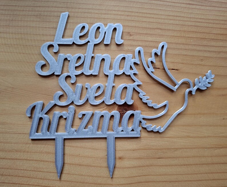 May include: A silver-colored cake topper featuring the text Leon Sretna Sveta Krizma in a cursive font, accompanied by a dove silhouette holding an olive branch. The topper has two pointed stakes at the bottom for insertion into a cake surface.