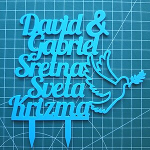 May include: A bright blue cake topper featuring the text "David & Gabriel Sretna Sveta Krizma" in a cursive font, accompanied by a dove silhouette holding an olive branch. The topper includes two pointed stakes at the base for insertion.