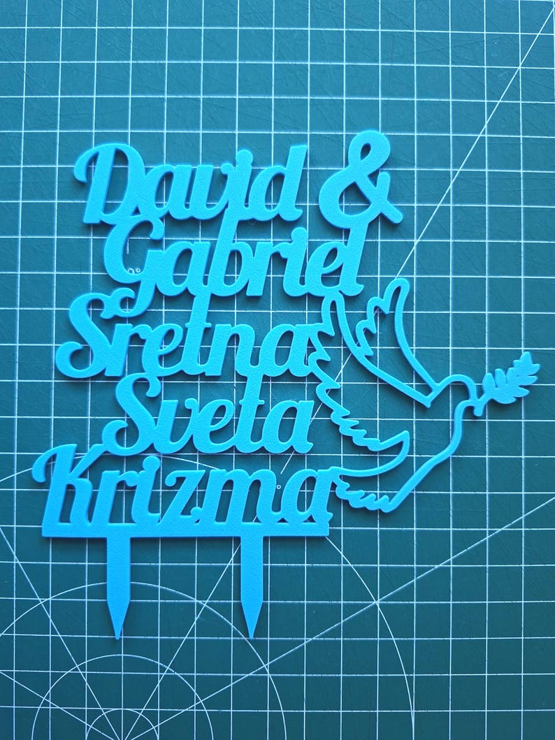 May include: A bright blue cake topper featuring the text "David & Gabriel Sretna Sveta Krizma" in a cursive font, accompanied by a dove silhouette holding an olive branch. The topper includes two pointed stakes at the base for insertion.