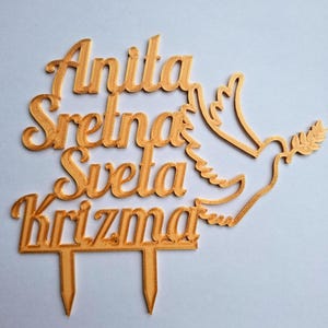 May include: A gold-colored cake topper featuring the text Anita Sretna Sveta Krizma in a cursive font, accompanied by a stylized dove carrying an olive branch. The topper includes two pointed stakes at the bottom for insertion into a cake.