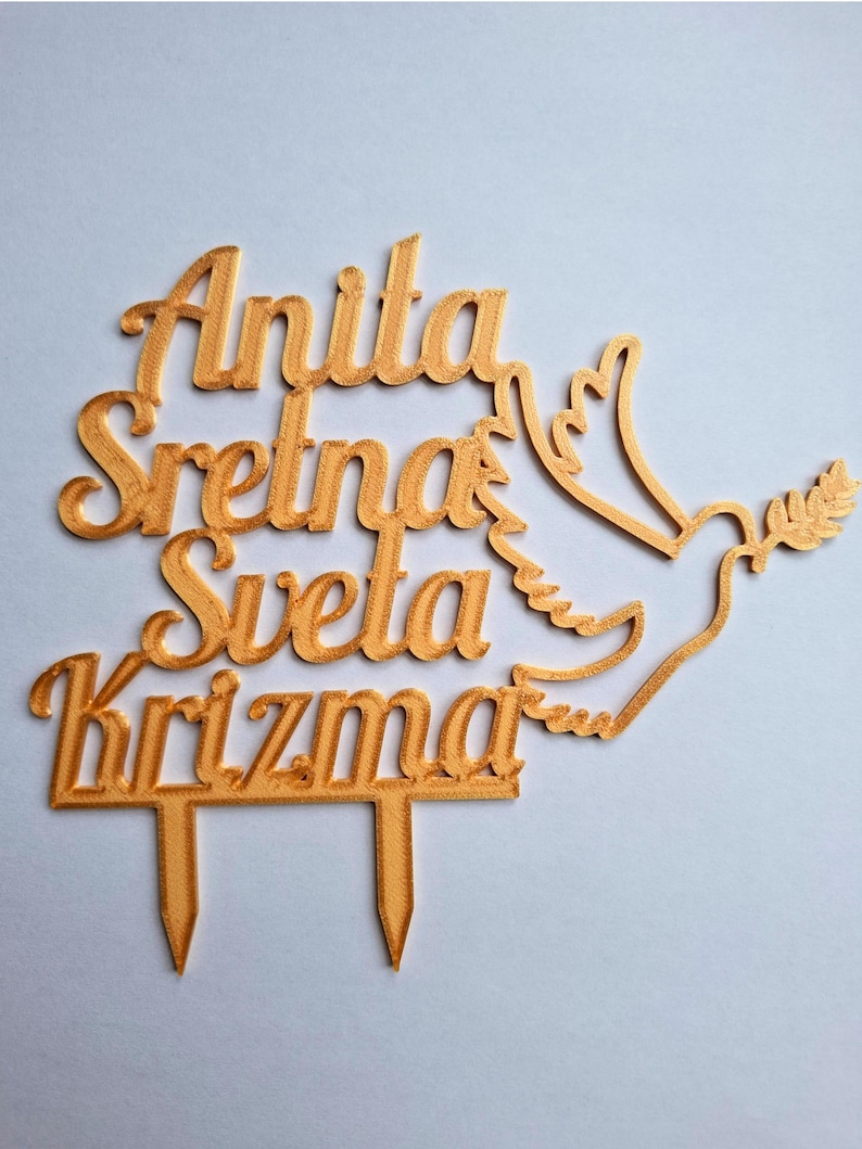 May include: A gold-colored cake topper featuring the text Anita Sretna Sveta Krizma in a cursive font, accompanied by a stylized dove carrying an olive branch. The topper includes two pointed stakes at the bottom for insertion into a cake.
