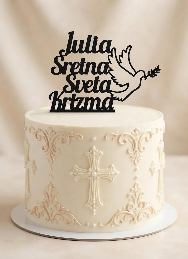 May include: A black cake topper featuring the text Julia Sretna Sveta Krizma and a dove holding an olive branch, positioned on a white cake decorated with cream-colored piped cross motifs and small pearl accents on the sides.