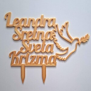 May include: A gold-colored cake topper featuring the text Leandra Sretna Sveta Krizma in a cursive font, accompanied by a stylized dove silhouette holding an olive branch. The topper includes two pointed stakes at the bottom for insertion.