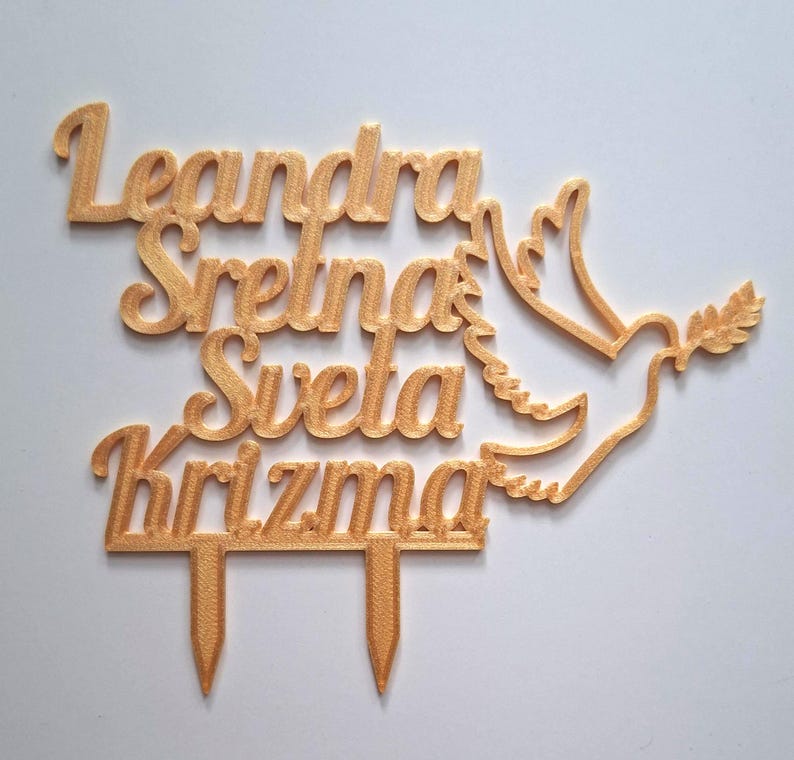 May include: A gold-colored cake topper featuring the text Leandra Sretna Sveta Krizma in a cursive font, accompanied by a stylized dove silhouette holding an olive branch. The topper includes two pointed stakes at the bottom for insertion.