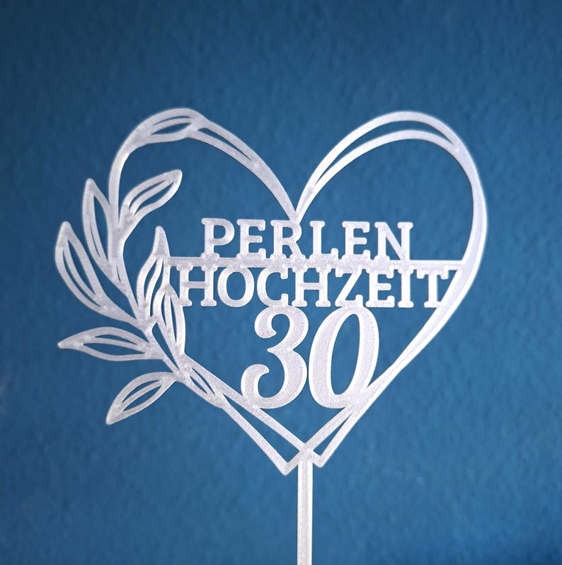 Może przedstawiać: Biały topper na tort w kształcie serca z gałązką liści po lewej stronie. Tekst PERLEN HOCHZEIT jest wyśrodkowany, a poniżej znajduje się liczba 30. Topper posiada pojedynczy pionowy patyczek u podstawy.