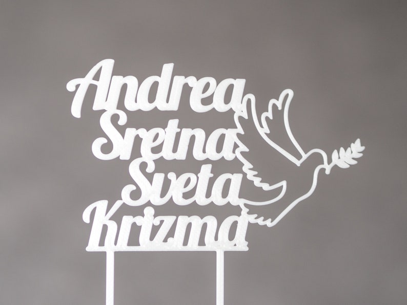 May include: A white cake topper featuring the text "Andrea Sretna Sveta Krizma" in a cursive font, accompanied by a silhouette of a dove holding an olive branch. The topper includes two vertical prongs at the base for insertion into a cake.