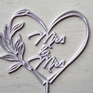 Op de afbeelding: Een zilverkleurige taarttopper met een hartvormig ontwerp met de woorden "Mrs & Mrs". De topper bevat een decoratieve tak en is geschikt voor een bruiloft of verlovingsceremonie.