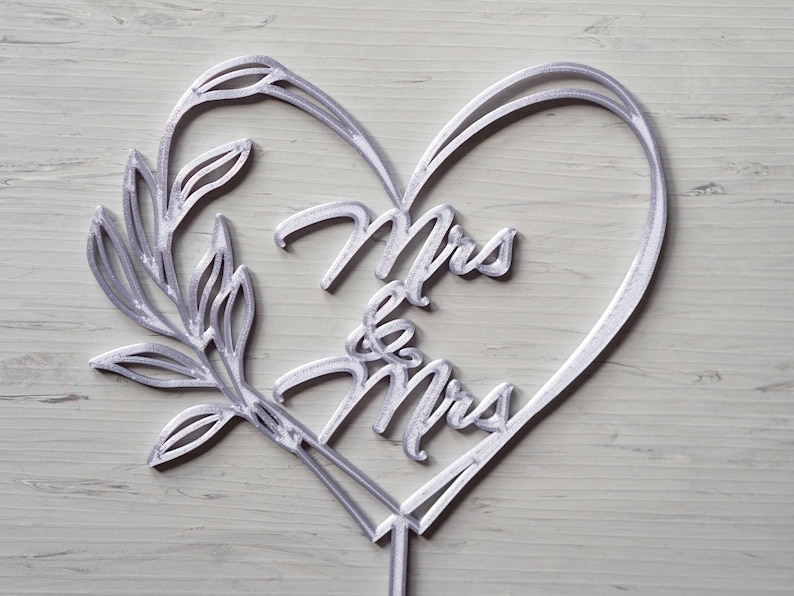 Op de afbeelding: Een zilverkleurige taarttopper met een hartvormig ontwerp met de woorden "Mrs & Mrs". De topper bevat een decoratieve tak en is geschikt voor een bruiloft of verlovingsceremonie.