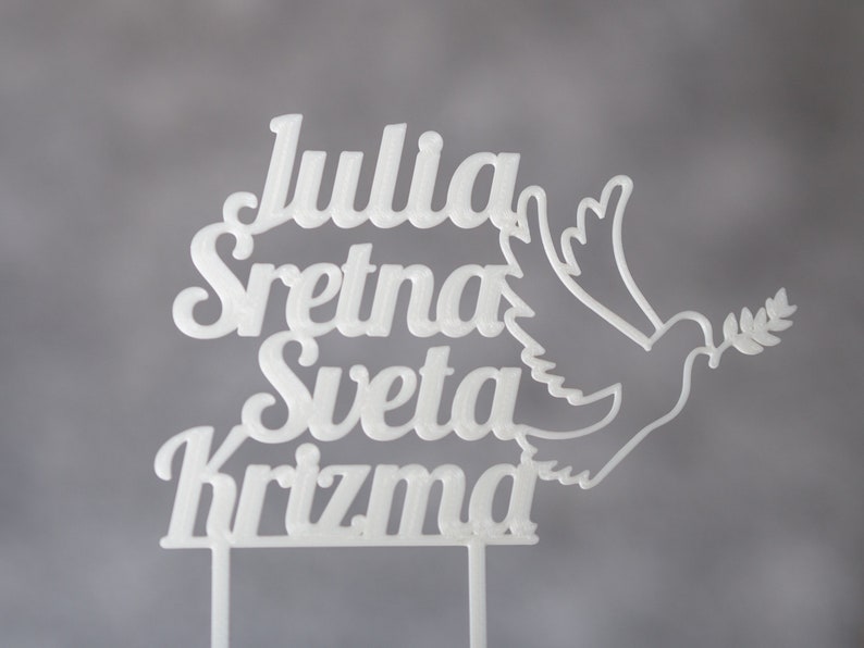 Könnte beinhalten: Ein weißer Tortenstecker mit dem Text Julia Sretna Sveta Krizma in einer stilisierten Schriftart, begleitet von einer Grafik einer Taube, die einen Olivenzweig hält. Der Tortenstecker hat zwei dünne vertikale Stäbe an der Basis.