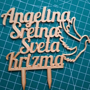 May include: A copper-colored cake topper featuring the text Angelina Sretna Sveta Krizma in a cursive font, accompanied by a dove silhouette holding an olive branch. The topper includes two pointed stakes at the bottom for insertion into a cake.