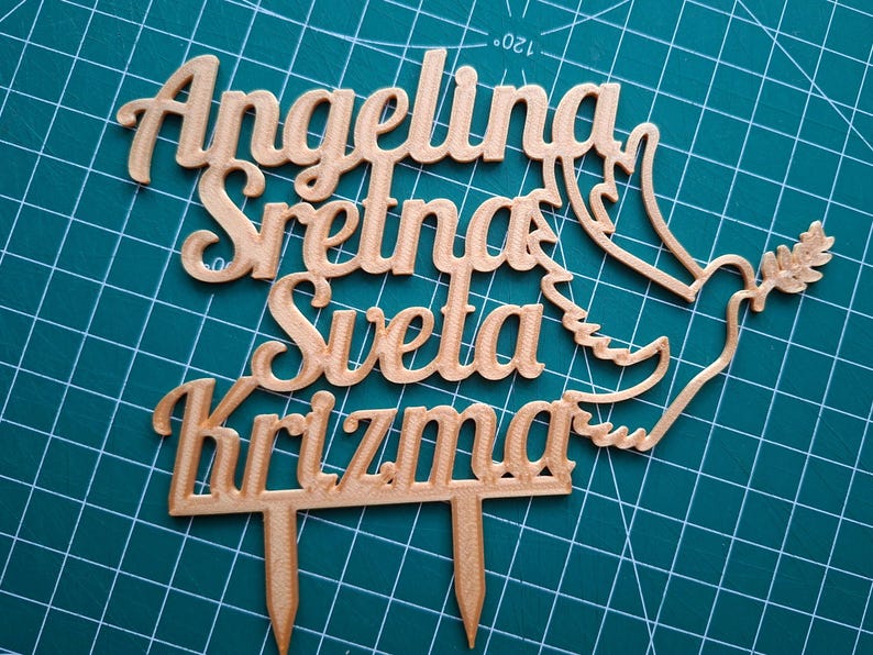 May include: A copper-colored cake topper featuring the text Angelina Sretna Sveta Krizma in a cursive font, accompanied by a dove silhouette holding an olive branch. The topper includes two pointed stakes at the bottom for insertion into a cake.