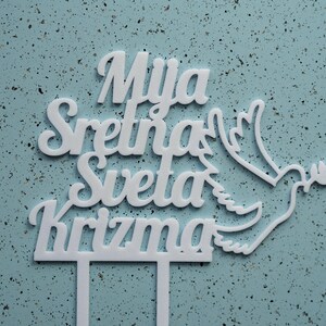 May include: A white cake topper featuring the text Mija Sretna Sveta Krizma in a cursive font, accompanied by a stylized dove graphic holding an olive branch. The topper includes two vertical prongs at the base for insertion into a cake surface.