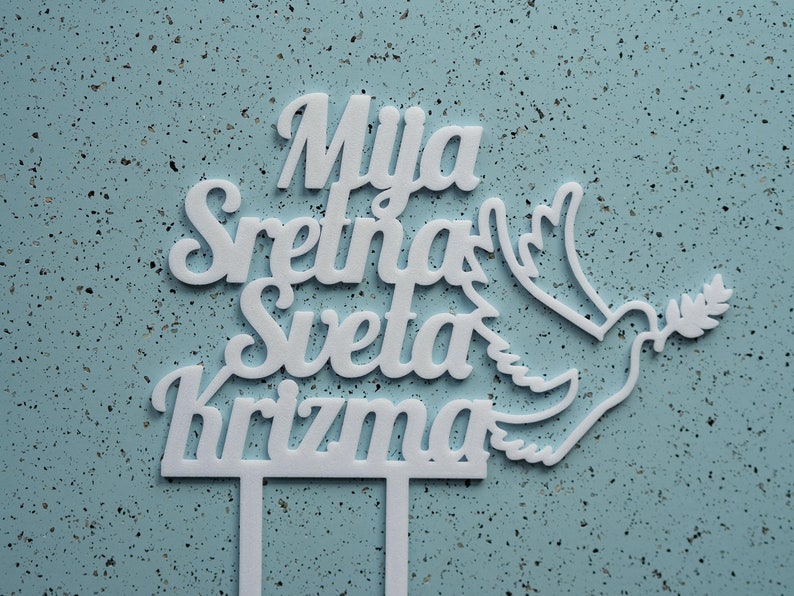May include: A white cake topper featuring the text Mija Sretna Sveta Krizma in a cursive font, accompanied by a stylized dove graphic holding an olive branch. The topper includes two vertical prongs at the base for insertion into a cake surface.