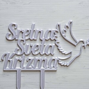 May include: A silver-colored cake topper featuring the text Sretna Sveta Krizma in a cursive font, accompanied by a stylized dove holding an olive branch. The topper includes two vertical prongs at the base for insertion into a cake surface.