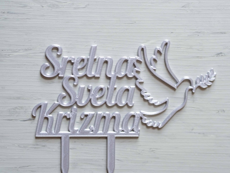 May include: A silver-colored cake topper featuring the text Sretna Sveta Krizma in a cursive font, accompanied by a stylized dove holding an olive branch. The topper includes two vertical prongs at the base for insertion into a cake surface.