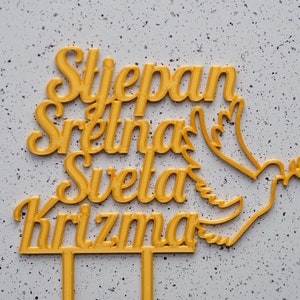 May include: A gold-colored cake topper featuring the text Stjepan Sretna Sveta Krizma in a cursive script font, accompanied by a dove silhouette holding an olive branch. The topper includes two vertical prongs at the base for insertion.