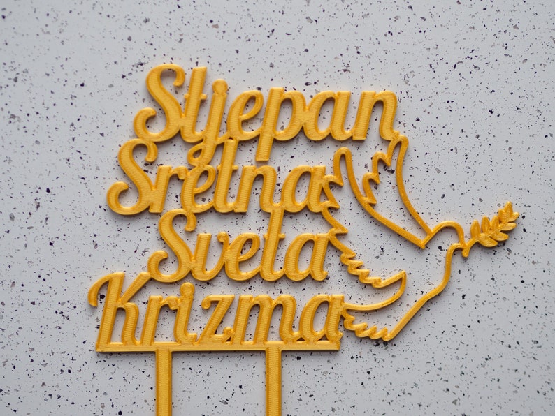 May include: A gold-colored cake topper featuring the text Stjepan Sretna Sveta Krizma in a cursive script font, accompanied by a dove silhouette holding an olive branch. The topper includes two vertical prongs at the base for insertion.